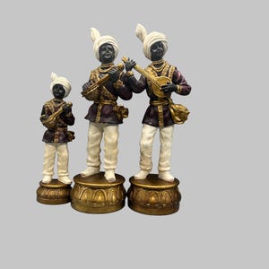 May include: Three decorative figurines of musicians. Each figure is dressed in a turban, white pants, and a dark purple jacket with gold accents. The musicians are playing instruments and standing on gold pedestals.