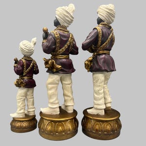 May include: Three decorative figurines of figures in traditional attire. The figures have black skin, white turbans, and are dressed in purple jackets with gold accents and white pants. They stand on ornate gold bases.