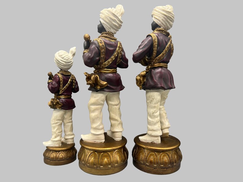 May include: Three decorative figurines of figures in traditional attire. The figures have black skin, white turbans, and are dressed in purple jackets with gold accents and white pants. They stand on ornate gold bases.