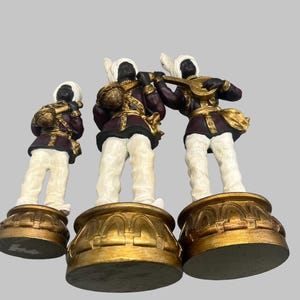 May include: Three decorative figurines of musicians. Each figure has a black face, white turban, and white pants. They wear burgundy jackets with gold accents and stand on gold bases. One figure plays a lute.
