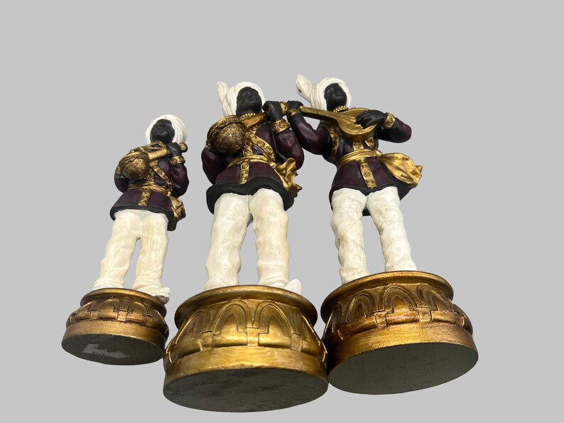 May include: Three decorative figurines of musicians. Each figure has a black face, white turban, and white pants. They wear burgundy jackets with gold accents and stand on gold bases. One figure plays a lute.