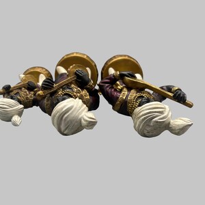 May include: Three decorative figurines of musicians. Each figure wears a turban, a purple and gold jacket, and holds a musical instrument. The figures are positioned on a light gray background.