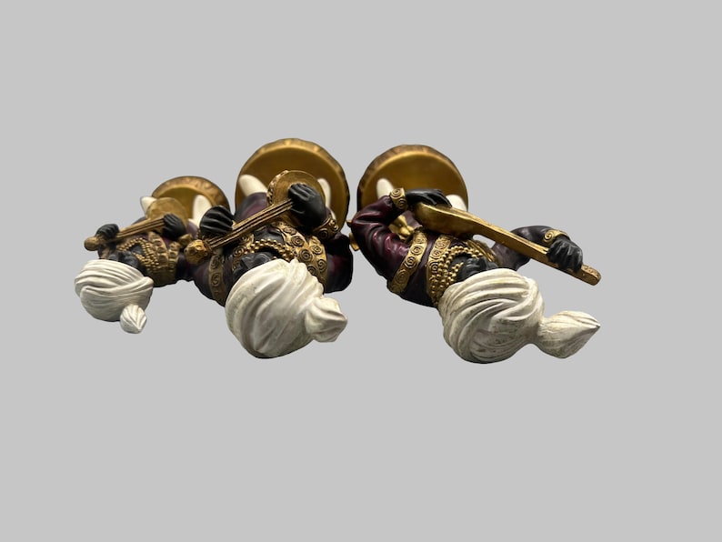 May include: Three decorative figurines of musicians. Each figure wears a turban, a purple and gold jacket, and holds a musical instrument. The figures are positioned on a light gray background.