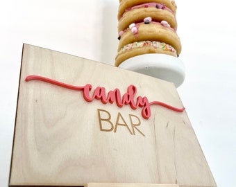 May include: A wooden "Candy Bar" sign with a pink cursive "candy" and gold "BAR" lettering, displayed on a small wooden easel. A stack of glazed donuts with sprinkles and marshmallows sits behind the sign on a white pedestal.