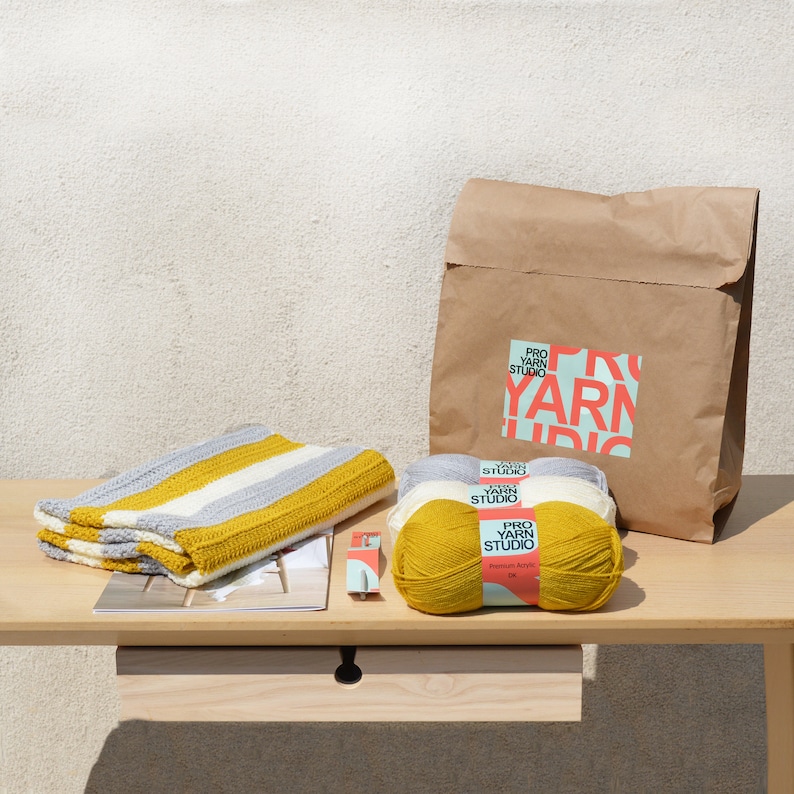 May include: A crocheted striped blanket in grey and yellow with three balls of yarn in white and yellow. The yarn is labeled "Pro Yarn Studio" and the bag is labeled "Pro Yarn Studio".
