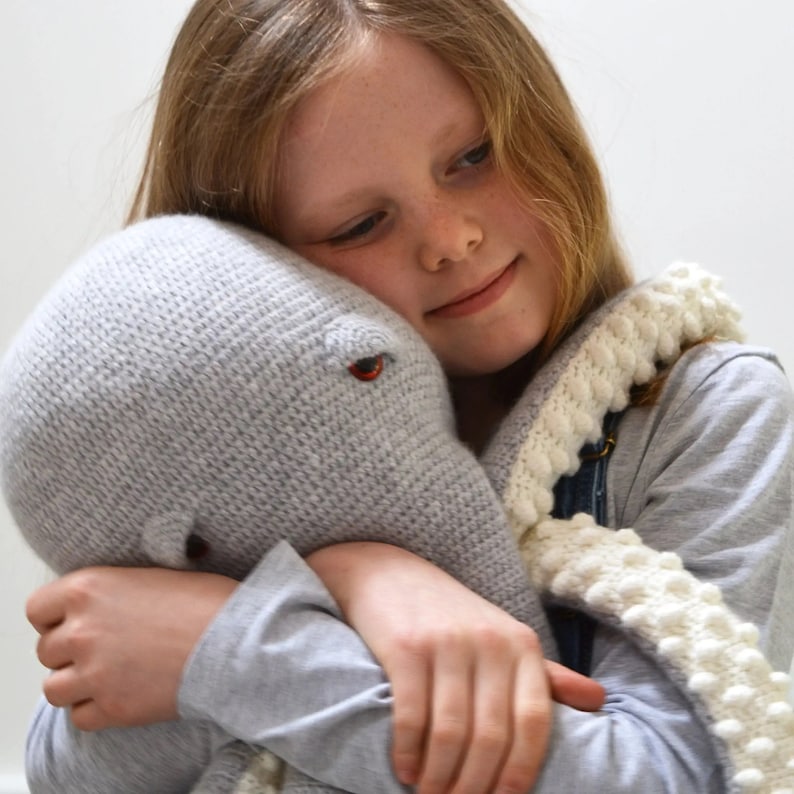 May include: A young girl hugs a gray crocheted octopus plush toy. The octopus has a white crocheted scarf with white pom-poms.