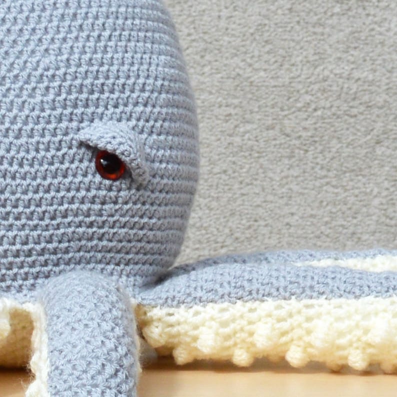 May include: A gray crocheted octopus with a white crocheted trim on the tentacles. The octopus has one red eye.