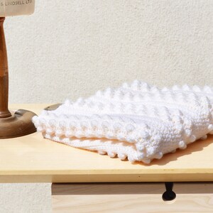 May include: A white crocheted baby blanket with a textured, puffy design. The blanket is folded and placed on a wooden surface.