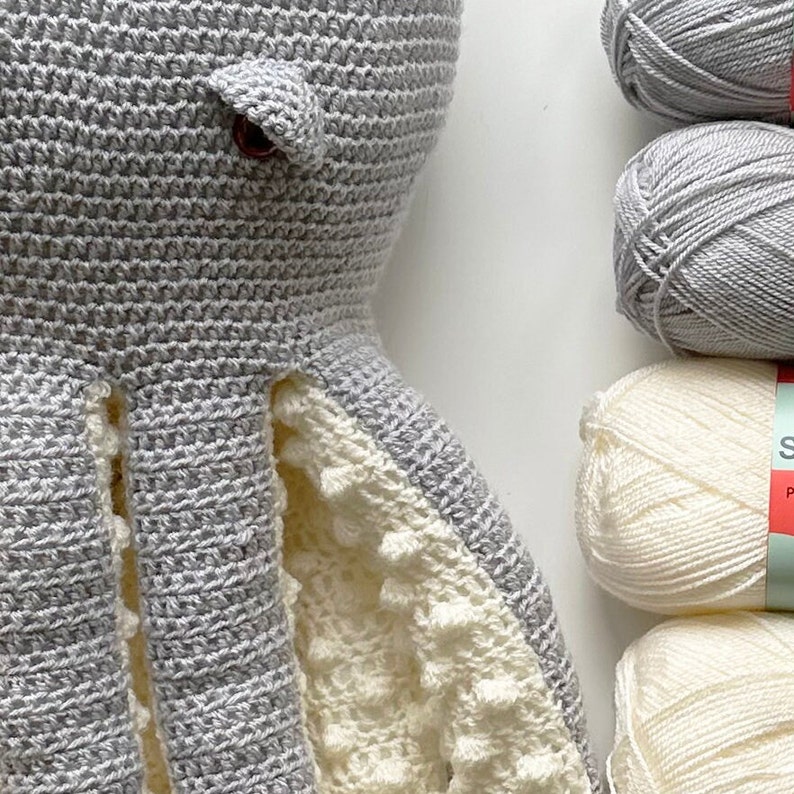May include: A gray crocheted octopus with white crocheted tentacles. The octopus has a brown button eye. The octopus is next to balls of gray and white yarn.