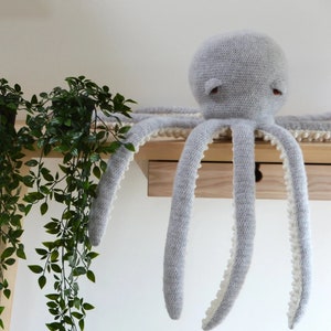 May include: A gray crocheted octopus with white trim around the edges of the tentacles. The octopus is sitting on a wooden shelf.
