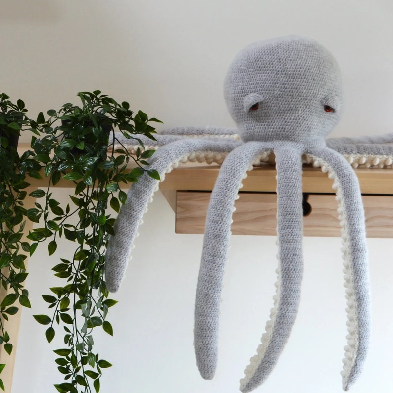 May include: A gray crocheted octopus with white trim around the edges of the tentacles. The octopus is sitting on a wooden shelf.