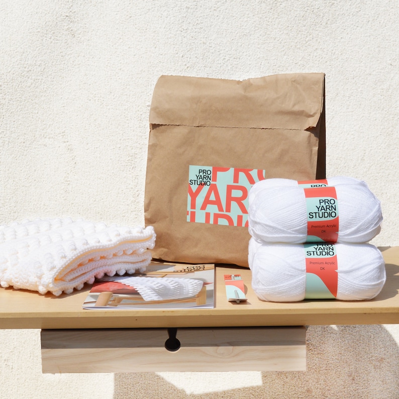 May include: A white crochet blanket with a brown paper bag and two white skeins of yarn labeled "Pro Yarn Studio" on a wooden table.