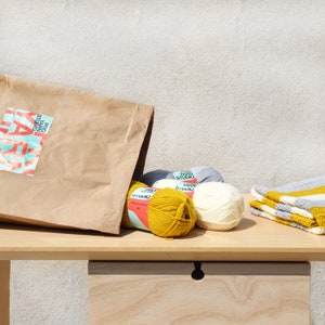 May include: A brown paper bag with the text "Pro Yarn Studio" on it, next to four balls of yarn in yellow, white, and gray. The yarn is on a wooden table with a drawer.