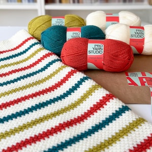 Op de afbeelding: Een gehaakt deken met strepen van wit, rood, groen en goud garen. Het deken is gemaakt met een eenvoudig steekpatroon. Vier bollen garen zijn zichtbaar op de achtergrond, met het label "Pro Yarn Studio" in rood en wit.