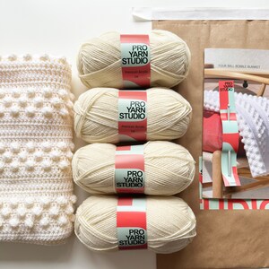May include: Four balls of cream-colored yarn labeled "Pro Yarn Studio" with the words "Premium Acrylic DK" on the label. The yarn is for crocheting a bobble blanket.