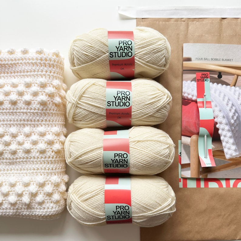 May include: Four balls of cream-colored yarn labeled "Pro Yarn Studio" with the words "Premium Acrylic DK" on the label. The yarn is for crocheting a bobble blanket.