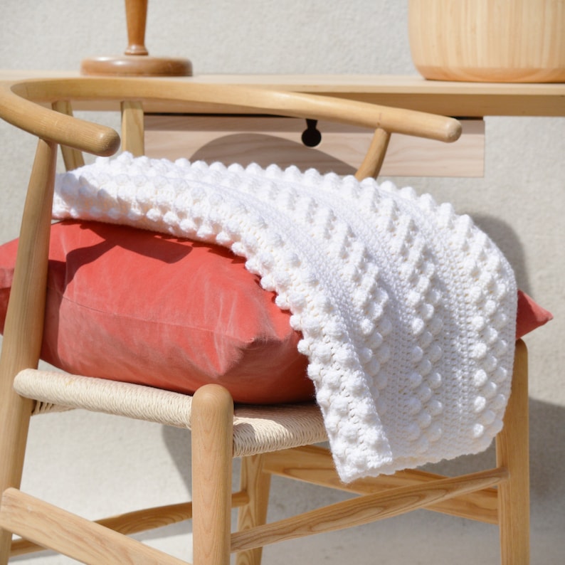 May include: A white crocheted throw blanket with a textured bobble stitch pattern draped over a wooden chair with a coral colored pillow.