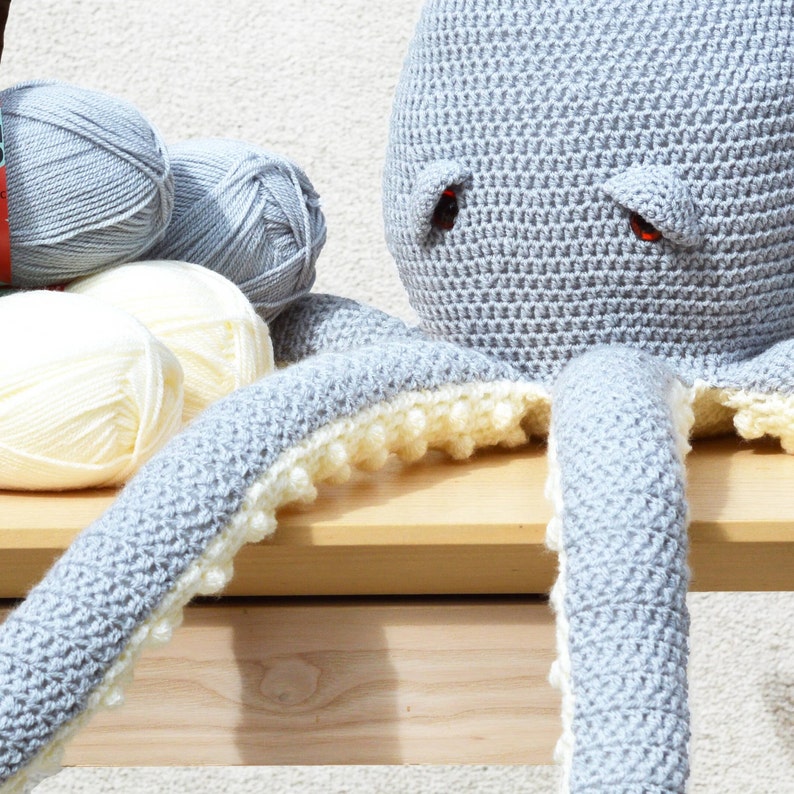 May include: A gray crocheted octopus with white crocheted accents. The octopus has large, red eyes and is sitting on a wooden surface.