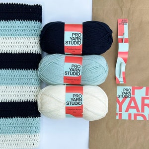 K&ouml;nnte beinhalten: Drei Kn&auml;uel Garn in Wei&szlig;, Hellblau und Dunkelblau mit Etiketten mit der Aufschrift "Pro Yarn Studio Premium Acrylic DK". Im Hintergrund befindet sich eine gestreifte H&auml;keldecke in Wei&szlig;, Hellblau und Dunkelblau.