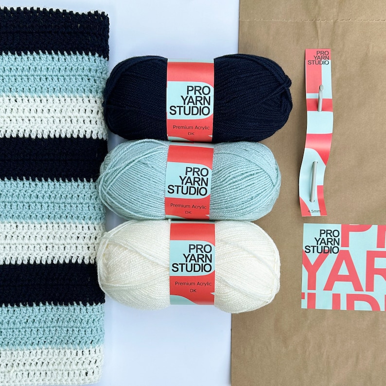 K&ouml;nnte beinhalten: Drei Kn&auml;uel Garn in Wei&szlig;, Hellblau und Dunkelblau mit Etiketten mit der Aufschrift "Pro Yarn Studio Premium Acrylic DK". Im Hintergrund befindet sich eine gestreifte H&auml;keldecke in Wei&szlig;, Hellblau und Dunkelblau.