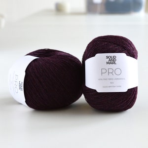 以下が含まれることがあります： ダークパープルの糸のボール2つ。ラベルには「Solid and Marl Pro 100% Fine Fibre Lambswool 50g Oiled British Yarn」と記載されています
