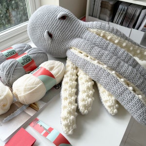 May include: A gray crocheted octopus with white crocheted tentacles. The octopus has a large head with a round, white eye. The octopus is lying on a white surface with two balls of yarn in the background.