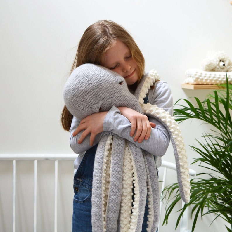 May include: A young girl hugs a gray and white crocheted octopus plush toy. The octopus has a long, curled tentacle and white pom-poms on the edges.