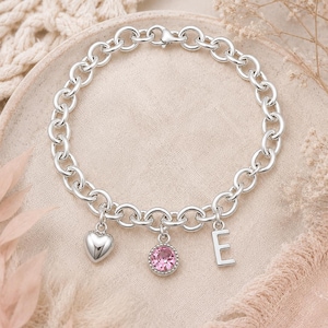 May include: A silver chain link bracelet featuring three dangling charms: a smooth polished heart, a round pink crystal set in a beaded silver bezel, and a capital letter E. The bracelet is secured with a lobster claw clasp closure.
