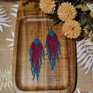 May include: A pair of long, beaded earrings with a teal and burgundy colour scheme. The earrings are hanging from a wooden tray.
