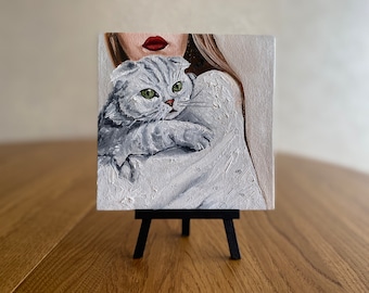 May include: A small painting on canvas of a woman with red lips holding a gray cat. The painting is on a small easel.