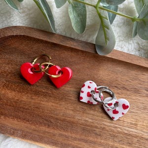 Peut inclure: Deux paires de boucles d'oreilles en forme de cœur. Une paire est rouge uni avec des cerceaux dorés, et l'autre est blanche avec un motif de cœur rouge et des cerceaux argentés. Les boucles d'oreilles sont présentées sur un plateau en bois avec de la verdure.