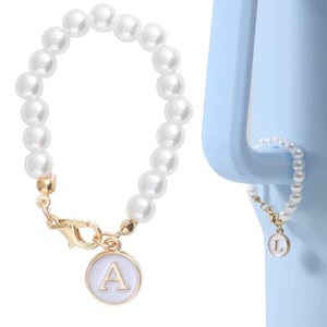 Peut inclure: Un bracelet de perles blanches avec un fermoir doré et un charm blanc avec la lettre "A". Un bracelet de perles plus petit avec un fermoir doré et un charm blanc avec la lettre "L" est également présenté. Les bracelets sont sur fond bleu clair.