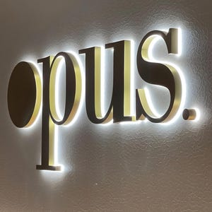 May include: Illuminated sign spelling "opus" in a modern font. The letters are a dark metallic color with a gold-colored interior and a bright white backlight, mounted on a textured gray wall. The sign is a decorative element.