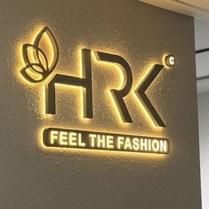 May include: Illuminated gold sign featuring the letters "HRK" and a butterfly motif. The text "FEEL THE FASHION" is displayed below. The sign is mounted on a textured, light-coloured wall.