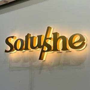 May include: A gold-coloured illuminated sign reading "Soluyshe" in a stylized font. The sign is mounted on a light grey wall. The letters are three-dimensional and have a warm, inviting glow.