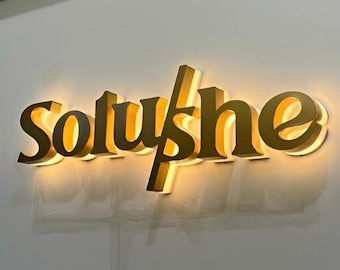 May include: A gold-colored illuminated sign that reads "Soluyshe" in a stylized font. The sign is mounted on a light gray wall. The letters are three-dimensional and have a warm, inviting glow.