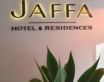 Może przedstawiać: Znak The JAFFA HOTEL & RESIDENCES z podświetlanymi czarnymi literami na białej ścianie. Litery mają żółtą poświatę. Zielone liście roślin na pierwszym planie.