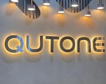May include: The word "QUTONE" is illuminated in a warm, golden light against a neutral-toned wall. Above, a series of pendant lights with gray shades and gold accents hang from the ceiling, casting soft light. The overall aesthetic is modern and inviting.
