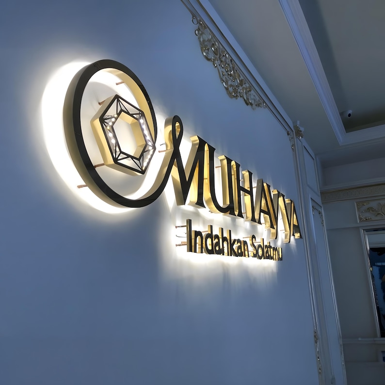 May include: A gold and black illuminated sign displaying the name "MUHAYA" and the text "Indahkan Solatmu". The sign incorporates a geometric design and is mounted on a light blue wall. The sign is lit with a warm glow.