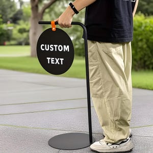 Könnte beinhalten: Ein schwarzes Metallschild mit rundem Anzeigebereich und gebogenem Griff. Auf dem Schild steht in weißer Schrift "CUSTOM TEXT". Ein orangefarbener Riemen befestigt das Schild am Griff. Das Schild steht auf einem schwarzen Sockel.