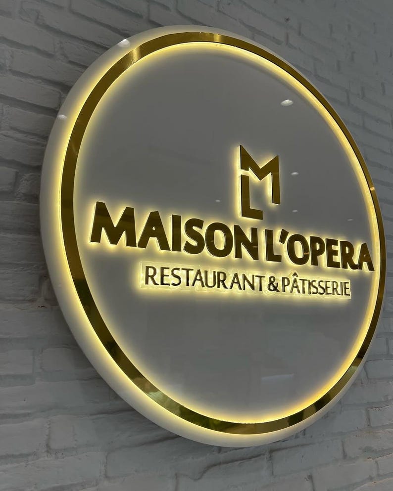 May include: A circular illuminated sign with the text "MAISON L'OPERA RESTAURANT & P&Acirc;TISSERIE" in gold. The sign has a gold border and a white background. It is mounted on a white brick wall.