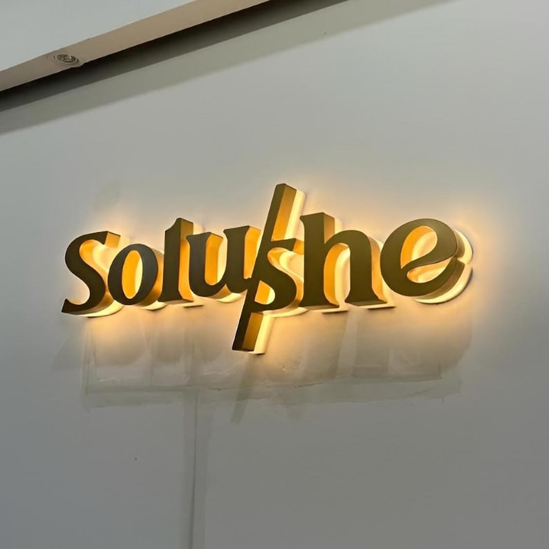 May include: A gold-coloured illuminated sign reading "Soluyshe" in a stylized font. The sign is mounted on a light grey wall. The letters are three-dimensional and have a warm, inviting glow.