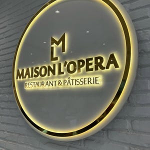 May include: A circular illuminated sign, featuring the words "MAISON L'OPERA RESTAURANT & P&Acirc;TISSERIE" in gold. The sign has a gold border and a white background, mounted on a white brick wall. The sign is round.