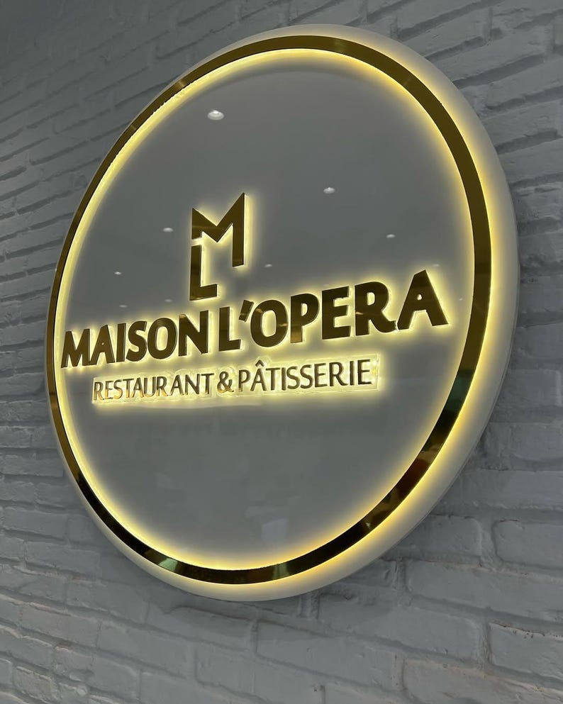 May include: A circular illuminated sign, featuring the words "MAISON L'OPERA RESTAURANT & P&Acirc;TISSERIE" in gold. The sign has a gold border and a white background, mounted on a white brick wall. The sign is round.