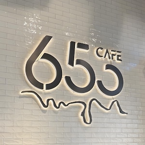 May include: A wall-mounted sign featuring the black numbers 655 and the word CAFE in black lettering. The sign is backlit, casting a soft glow against the white, rectangular, glossy subway-style tiles that form the background of the display.
