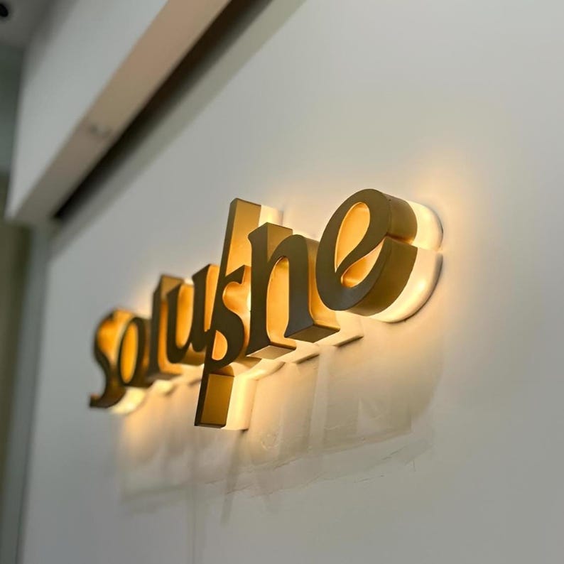 May include: A wall-mounted sign spelling "soushe" in gold, three-dimensional letters. The letters are backlit, producing a warm glow and a shadow on the white wall. The sign is likely for a business, possibly a shop or restaurant.