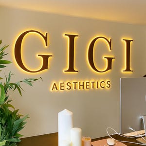 May include: A wall sign displaying the name "GIGI" in large, illuminated, brown letters, with "AESTHETICS" below in smaller, brown letters. The sign is lit with a warm, yellow glow, creating a welcoming atmosphere.