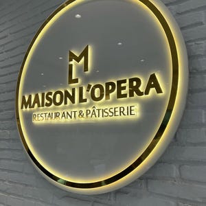 May include: Circular sign for "MAISON L'OPERA RESTAURANT & P&Acirc;TISSERIE" with a gold border and lettering. The sign has a white background and is mounted on a white brick wall. The logo includes the letters "M" and "L" in gold.