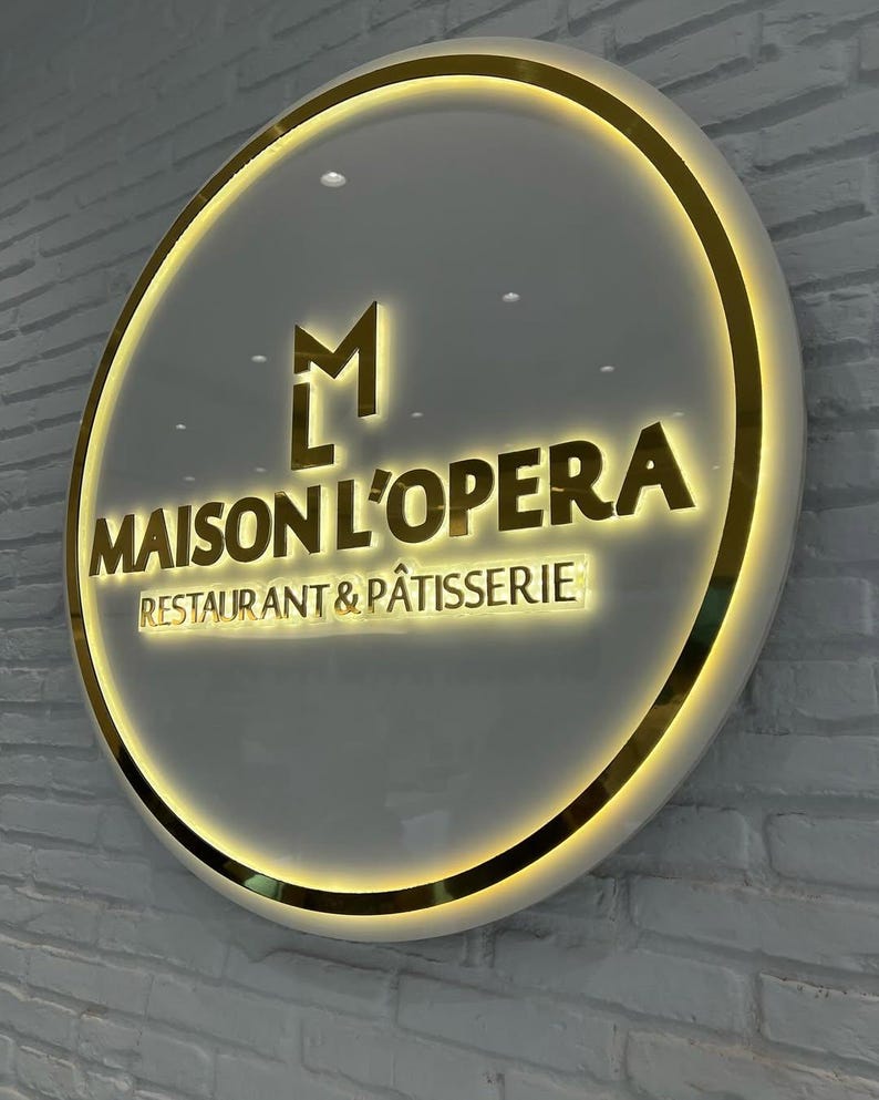 May include: Circular sign for "MAISON L'OPERA RESTAURANT & P&Acirc;TISSERIE" with a gold border and lettering. The sign has a white background and is mounted on a white brick wall. The logo includes the letters "M" and "L" in gold.