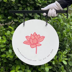 Puede incluir: Un letrero redondo blanco con una ilustración de flor de loto rosa y el texto "www.buddhablissSF.com Tel. 415. 565. 9634". El letrero está suspendido por cadenas de un soporte de metal negro.